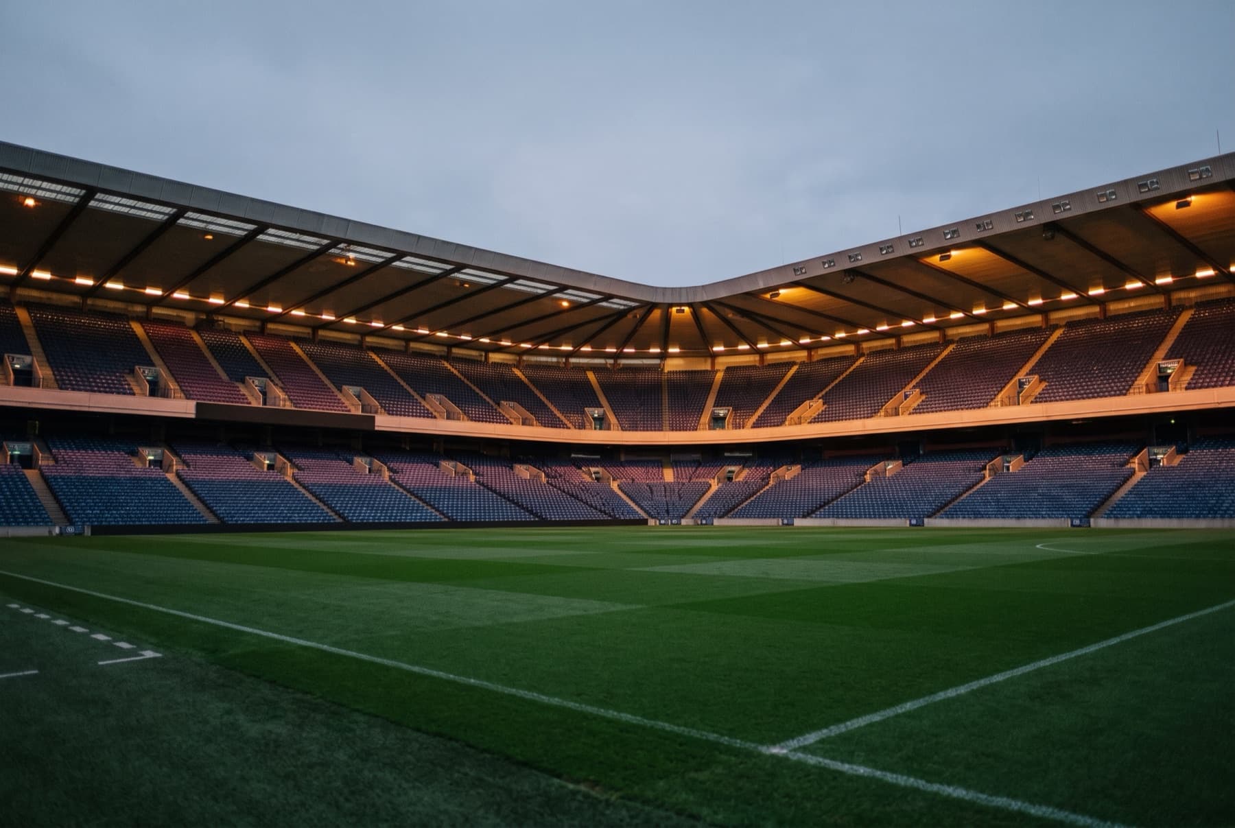 An empty top-flight football stadium at dusk, floodlights on, three tiers of seats